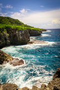 A high-angle view of rugged, porous limestone cliffs and a natural stone arch on the eastern coast of Guam, with blue Pacific Ocean waves crashing against the rocks.