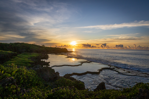 "A serene sunrise over the Inalåjan Natural Pools in Guam. The golden sun sits low on the horizon, casting a perfect mirror reflection onto the still, turquoise tide pools and weathered limestone karst formations."