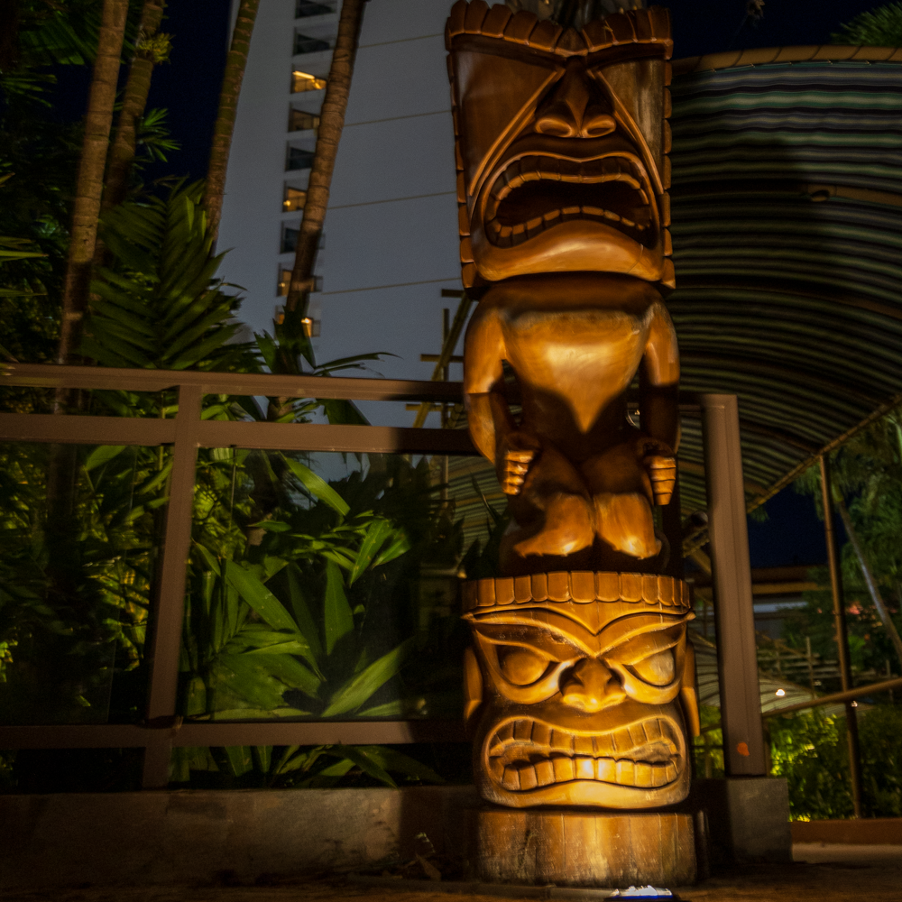 A close-up detail shot highlighting the warm uplighting, expressive face carvings, and polished wood grain of a wooden tiki totem.