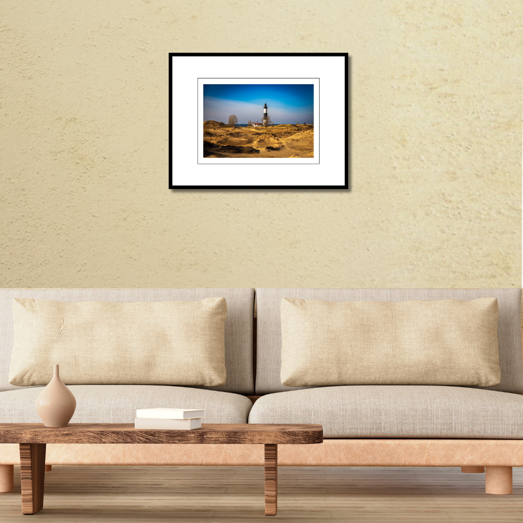 A framed landscape photography print of Big Sable Point Lighthouse displayed on a modern living room wall over a couch.