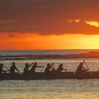 A close-up detail highlighting the sharp silhouettes of the canoe paddlers against a warm, glowing orange sky and horizon.