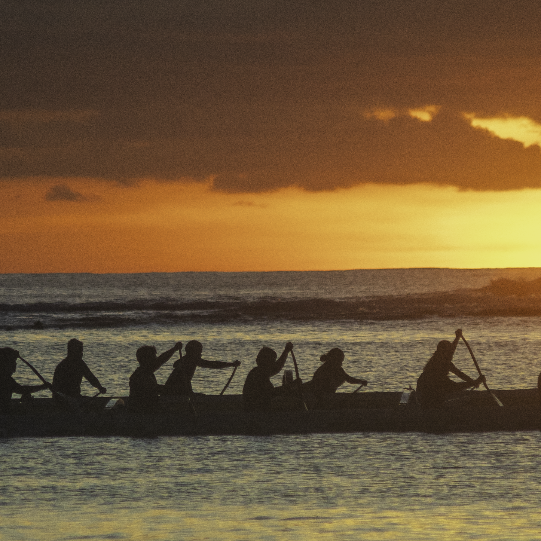 A close-up detail highlighting the sharp silhouettes of the canoe paddlers against a warm, glowing orange sky and horizon.