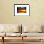 A framed coastal fine art print of an outrigger canoe silhouette at sunset displayed on a modern living room wall over a couch.