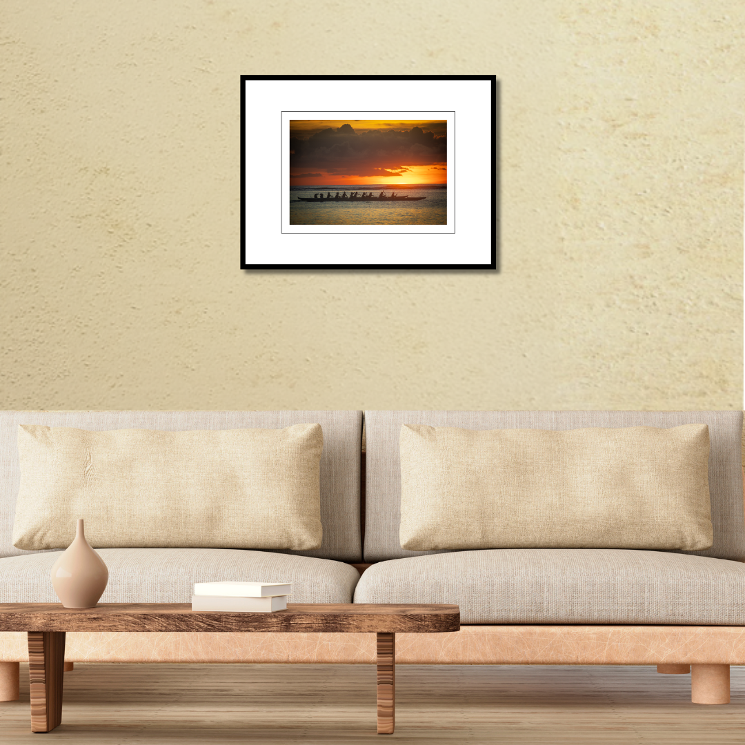 A framed coastal fine art print of an outrigger canoe silhouette at sunset displayed on a modern living room wall over a couch.