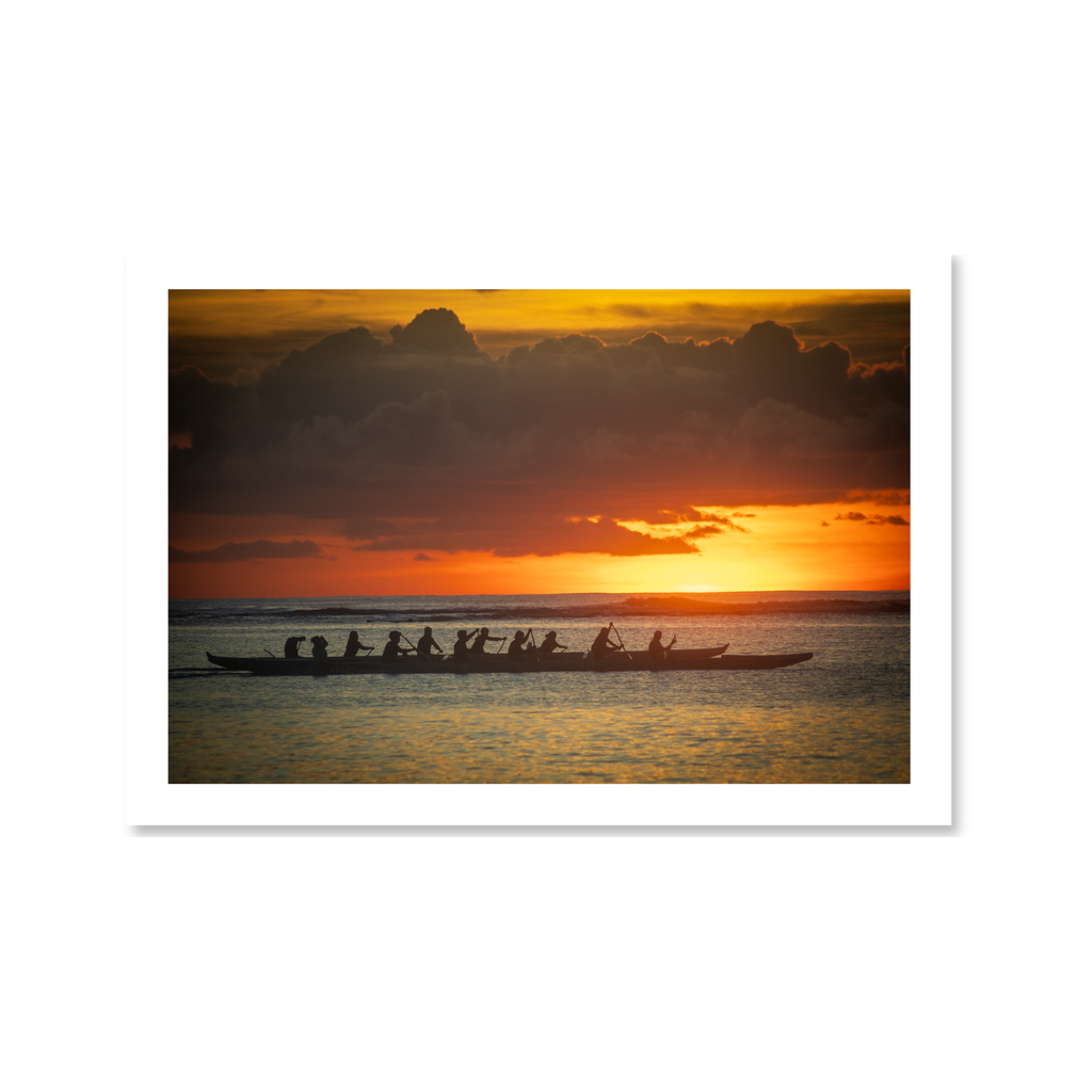 A striking silhouette of a traditional outrigger canoe team paddling across calm ocean waters during a vibrant golden sunset.