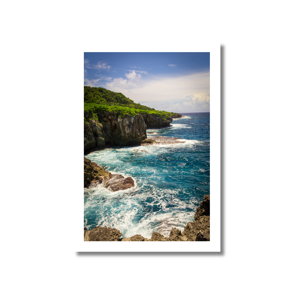 A high-angle view of rugged, porous limestone cliffs and a natural stone arch on the eastern coast of Guam, with blue Pacific Ocean waves crashing against the rocks.