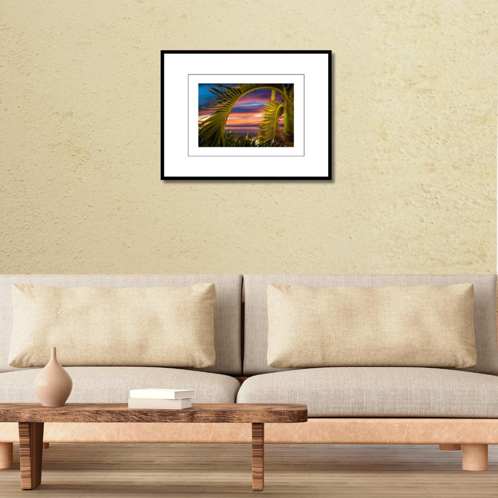 A framed tropical fine art landscape print of a palm tree sunset displayed on a modern wall over a couch to demonstrate scale.