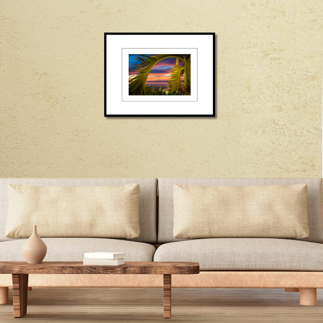 A framed tropical fine art landscape print of a palm tree sunset displayed on a modern wall over a couch to demonstrate scale.