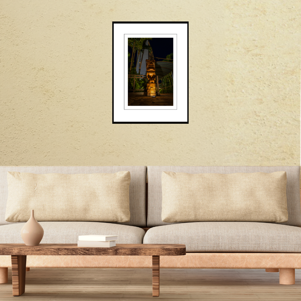 A framed fine art travel print of a carved tiki totem at night displayed on a living room wall above a modern couch to show scale.