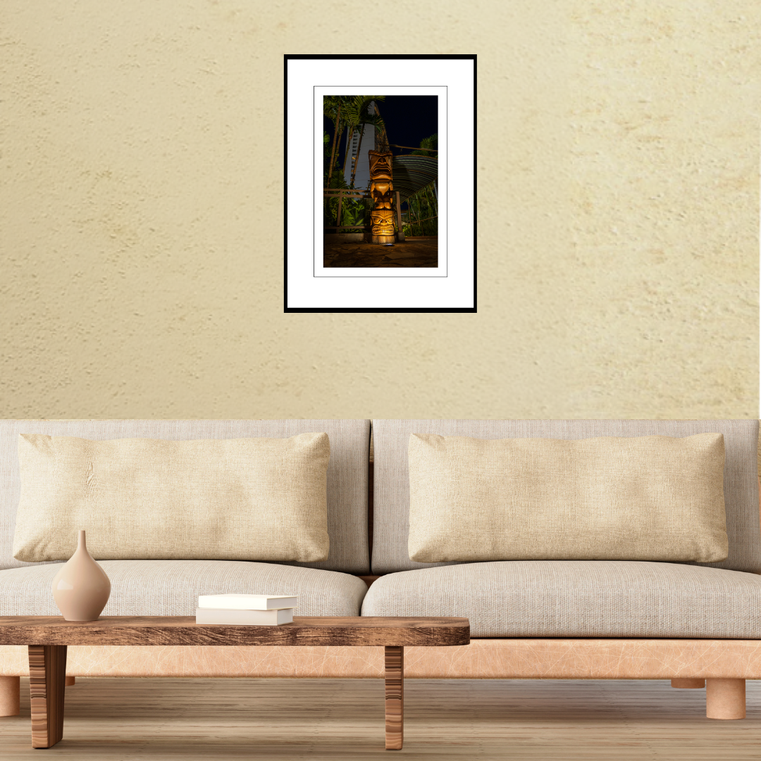 A framed fine art travel print of a carved tiki totem at night displayed on a living room wall above a modern couch to show scale.